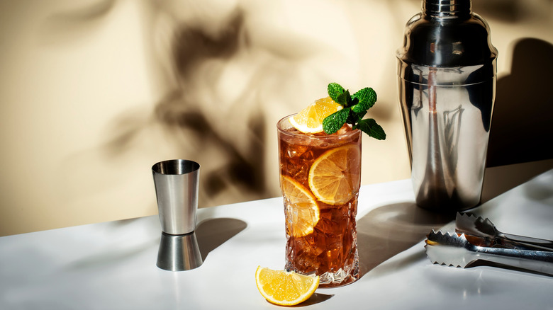 Long Island Iced tea garnished with mint and lemon, flanked by shaker, ice tongs, and shot measuring cup